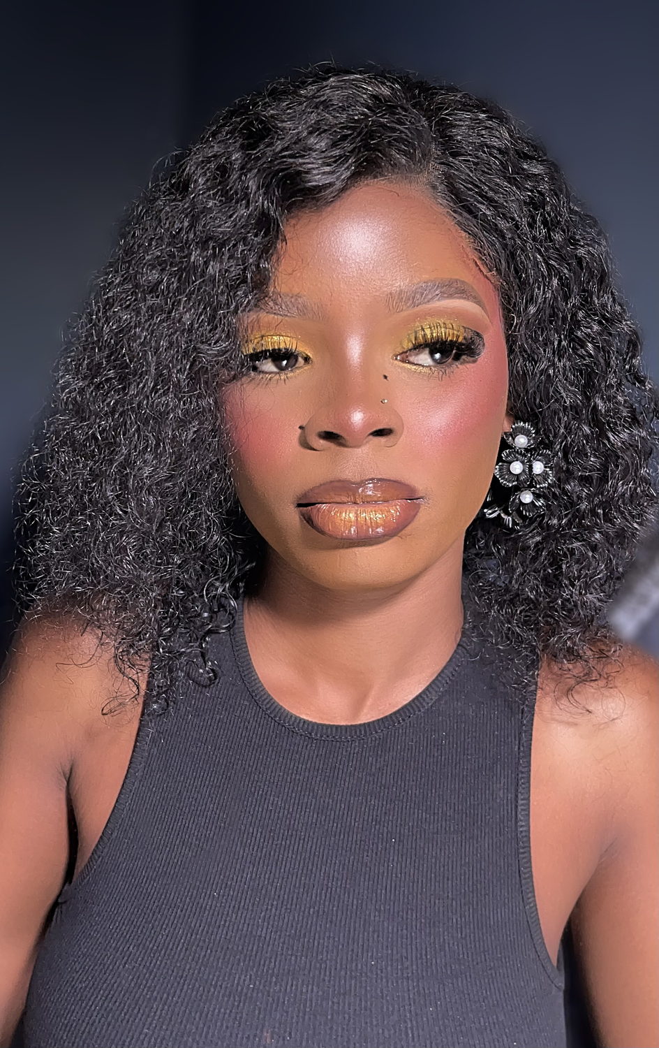 Gold shimmer eyeshadow with coral blush and a brown-to-gold ombre glossy lip, styled with wet-look curls and black floral earrings